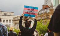 Person facing back at a protest holding a sign that says "Trans Folx Are Not A Threat, Transphobia Is"