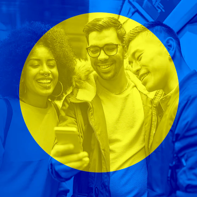 A Black woman, latinx man, and asian man look together, smiling, at the center man's phone.