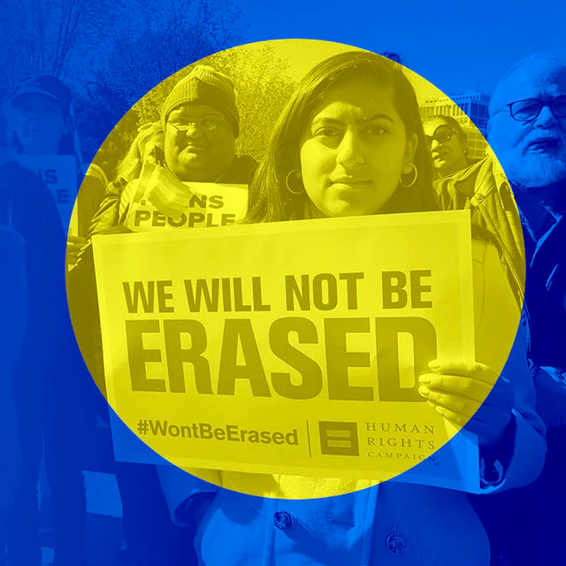 In a crowd of people, a trans woman holds a sign that reads "We Will Not Be Erased".
