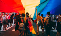 Crowd of people walking below a large billowing LGBTQ+ pride flag.